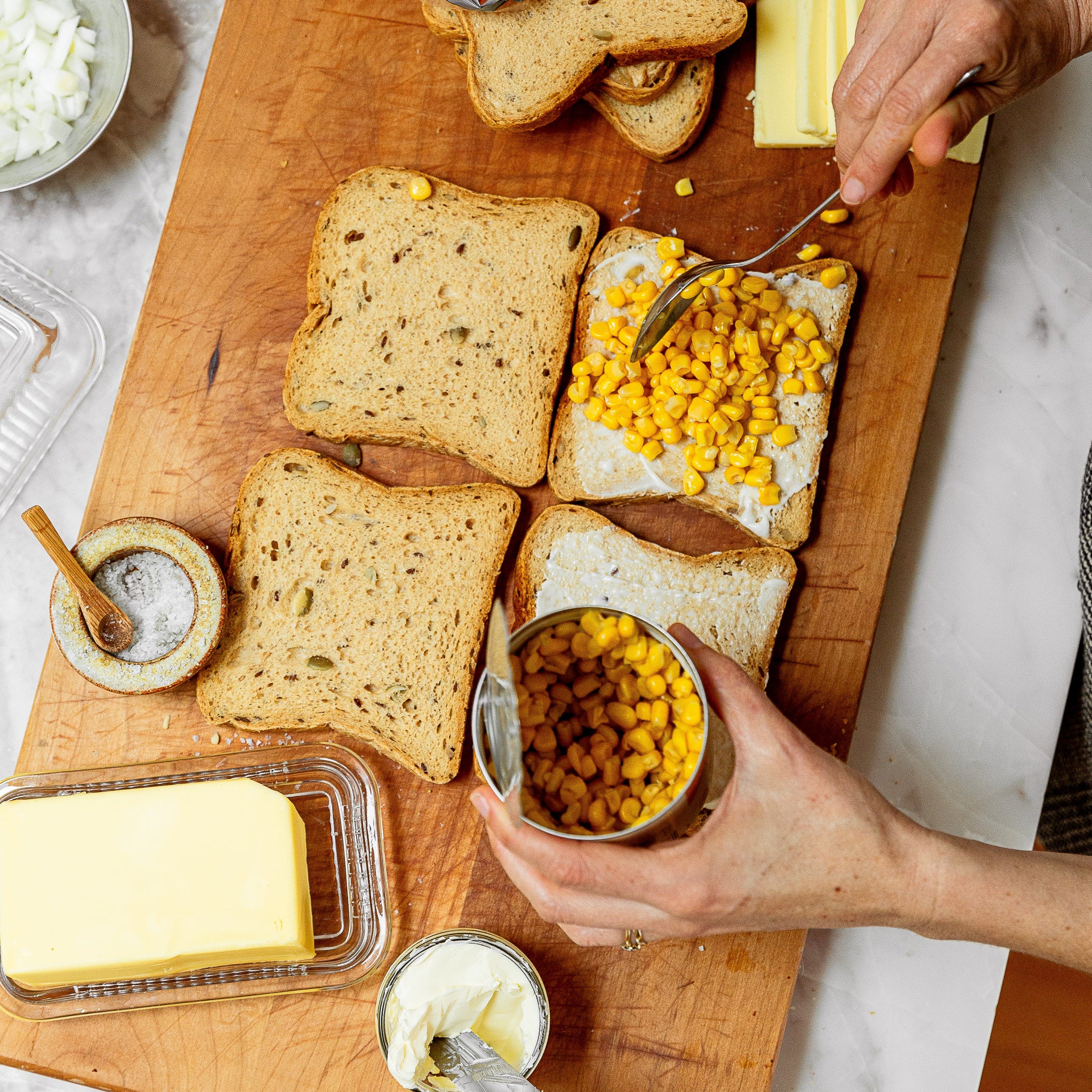 Overhead view of a wooden cutting board with slices of bread, butter, mayonnaise, and a can of corn kernels being spread onto a slice of bread by hand.