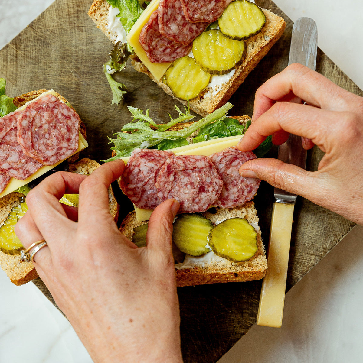 Hands assembling gourmet salami and cheese sandwiches with pickle slices on rustic bread, arranged on a wooden cutting board with a knife and a jar of pickles.