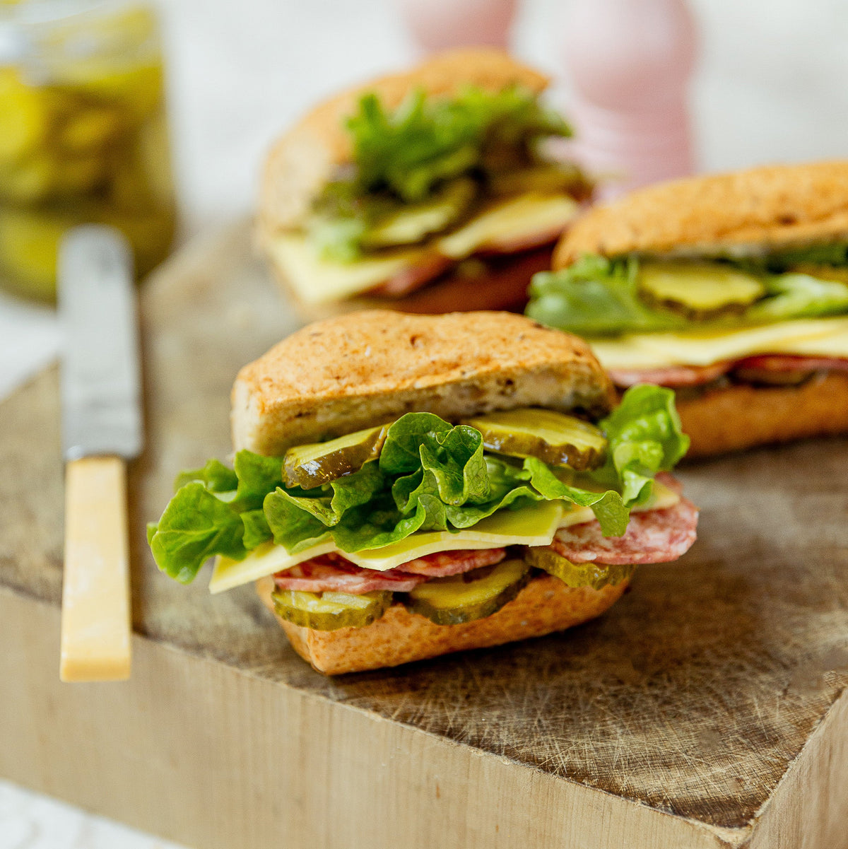 A close-up of a gourmet sandwich on a rustic wooden cutting board, featuring a multigrain bread bun, fresh green lettuce, pickles, and sliced salami, with two more sandwiches blurred in the background.