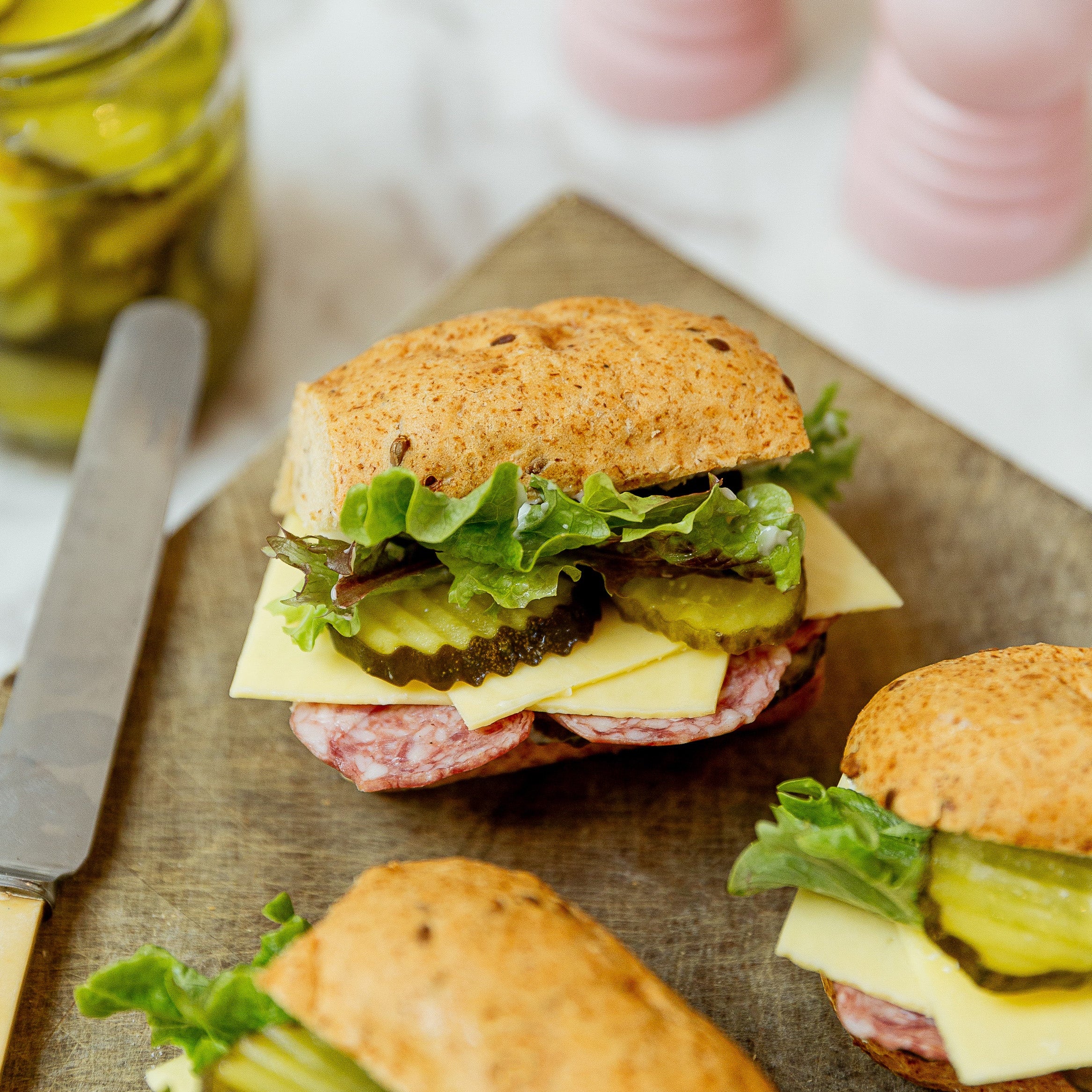 Seeded buns filled with salami, lettuce, pickles and cheese on a wooden chopping board. Pink salt and pepper grinders in the back with a jar of pickles and a butter knife.