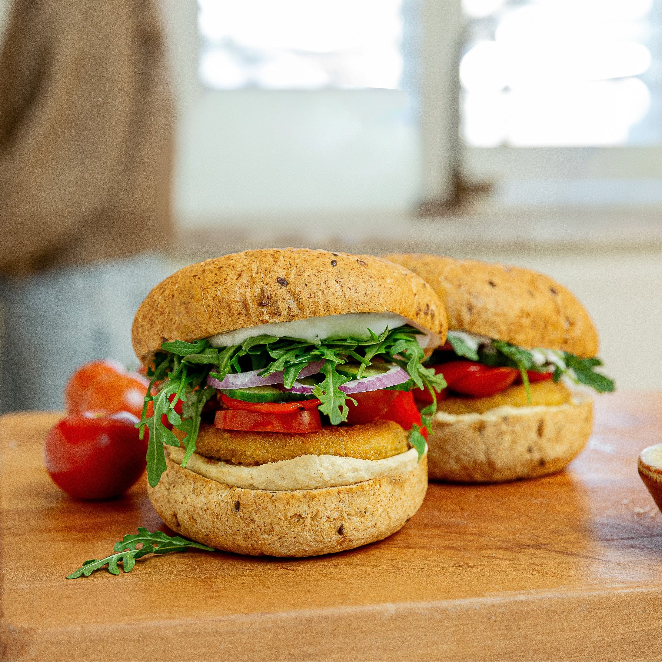 Two gourmet seeded buns filled with greens, tomatoes, and red onion, served on a wooden cutting board with fresh tomatoes and a small bowl of salt in a bright kitchen setting.