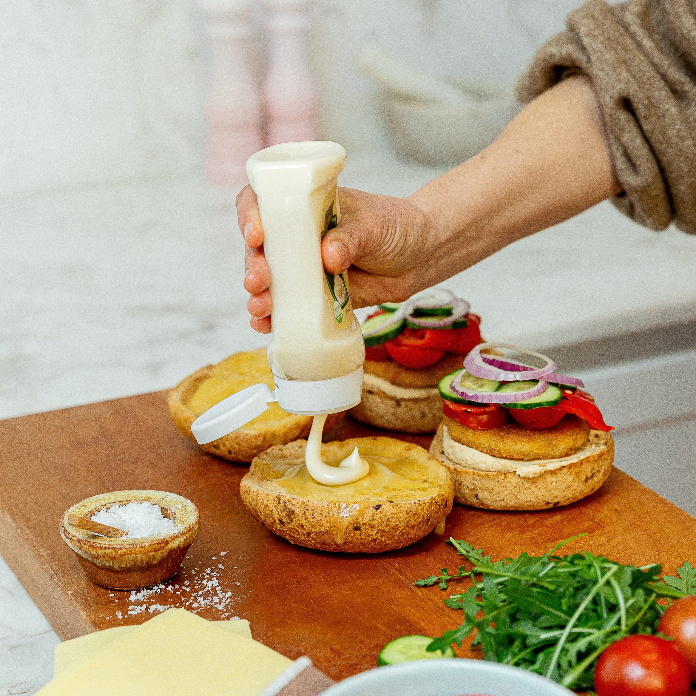 A close-up of a hand squeezing a bottle of creamy mayo onto burger buns on a wooden cutting board, with tomatoes, cucumbers, onions, cheese, and greens nearby in a bright kitchen setting.