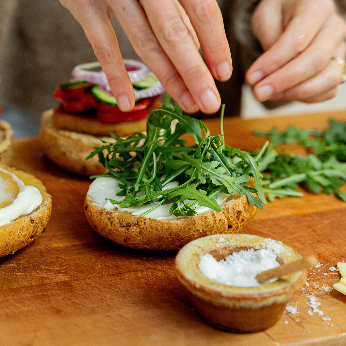 A close-up of a person's hands preparing a burger on a wooden cutting board, adding fresh arugula to a bagel with white spread, with other ingredients and small bowls of salt in the background.