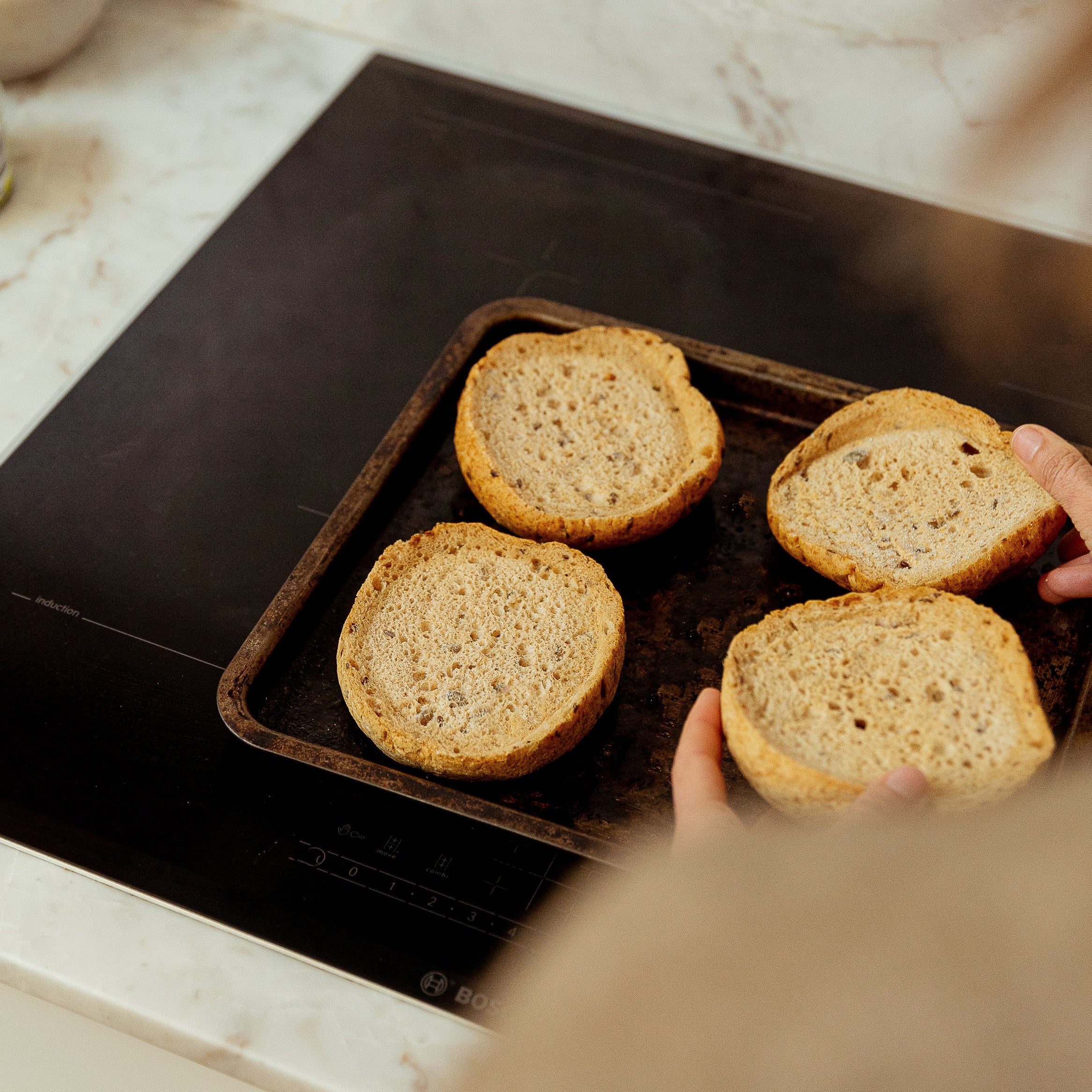 Four slices of toasted seeded buns on a dark baking tray, being held by hands in a kitchen setting with a marble countertop and black induction cooktop in the background.