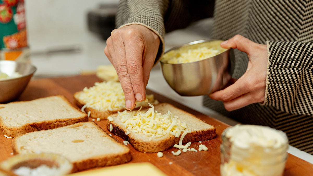 A close-up lifestyle shot of a person spreading shredded cheese onto slices of bread on a wooden cutting board, with a jar of butter and other baking ingredients nearby.