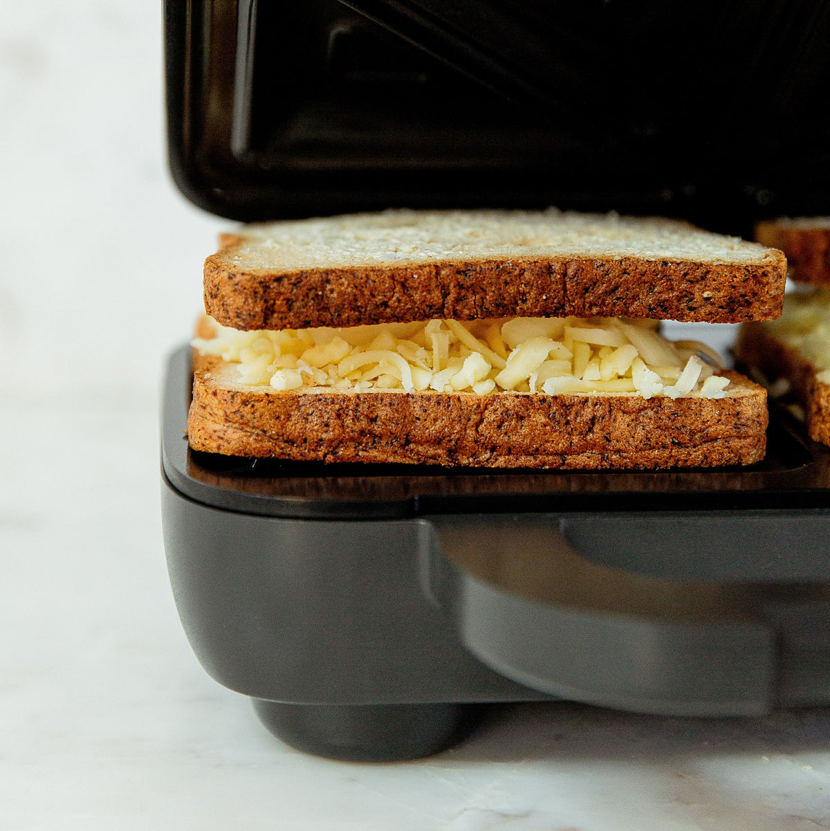 A close-up of a black sandwich press with a toasted sandwich in progress, featuring browned bread slices and melted cheese filling, set against a light marble background.