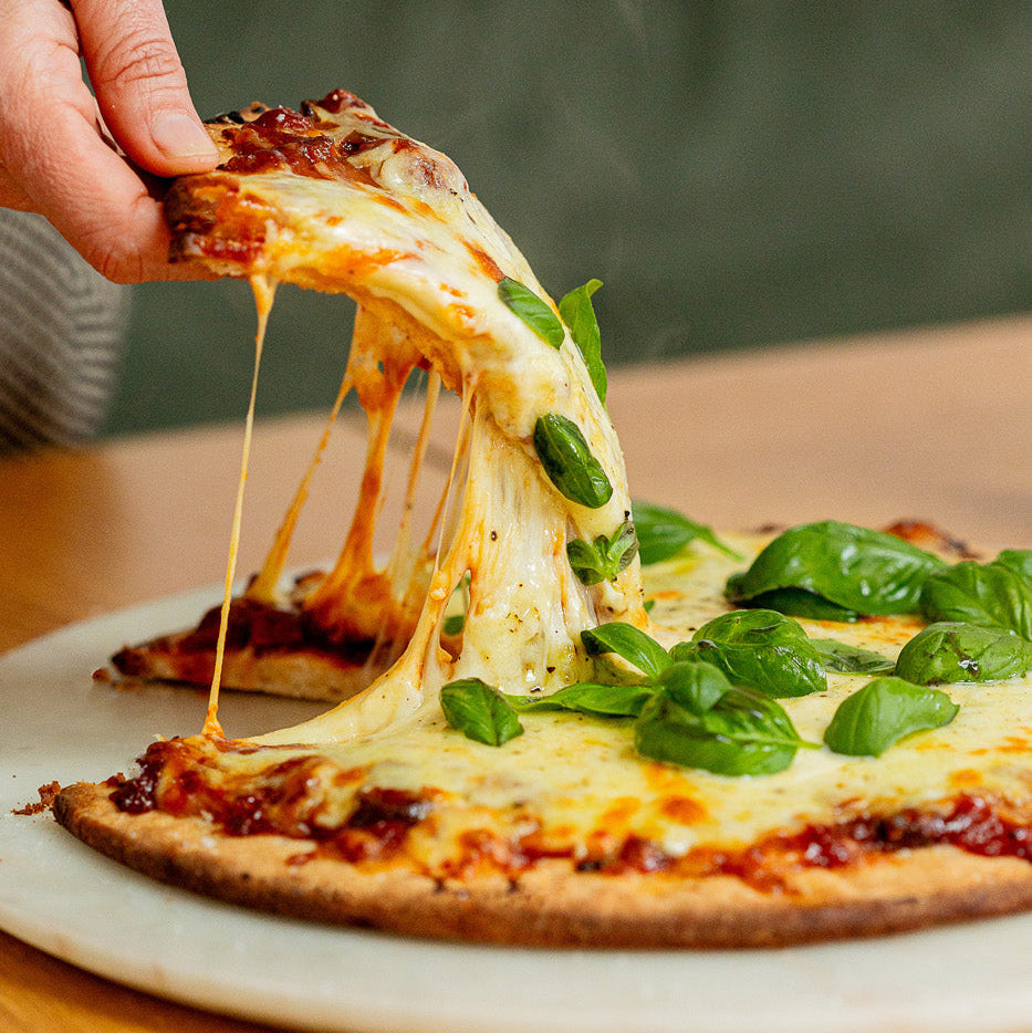 A close-up of a hand lifting a slice of cheesy pizza with basil from a round pizza on a white plate, set on a wooden table.