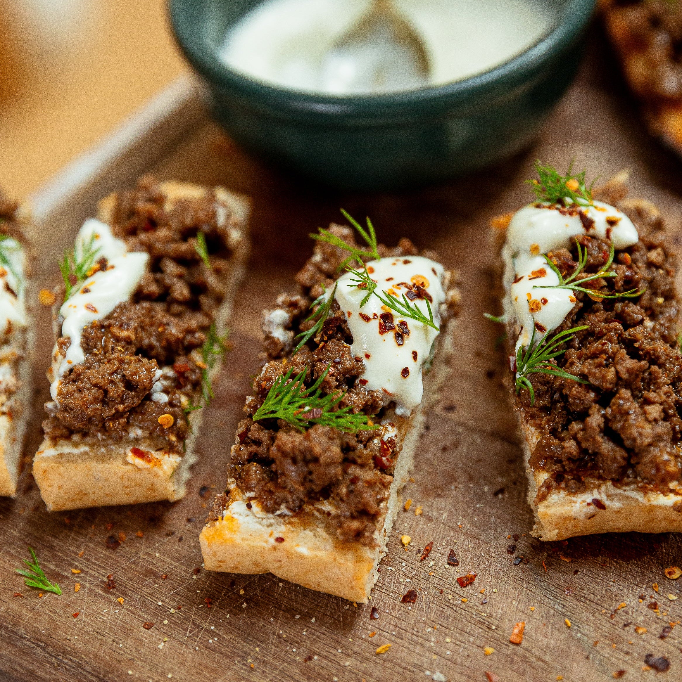 Sliced toasted bread topped with crumbled sausage, a dollop of white sauce, and fresh dill, arranged on a wooden board with a small bowl of white sauce and a spoon in the background.