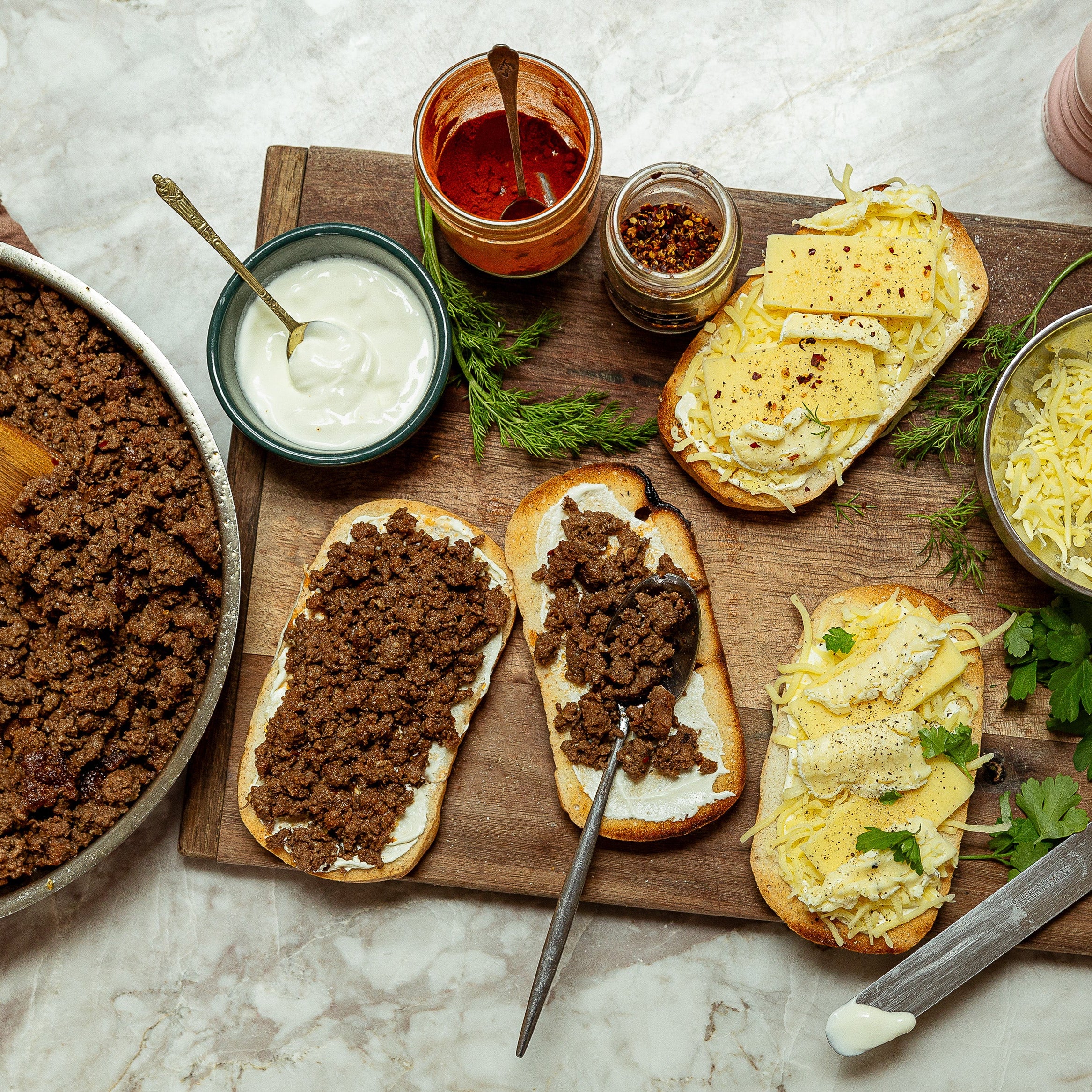 Overhead flat-lay of prepared gourmet Turkish pides on a wooden cutting board, surrounded by condiments, cheese, and spices on a light marble surface.