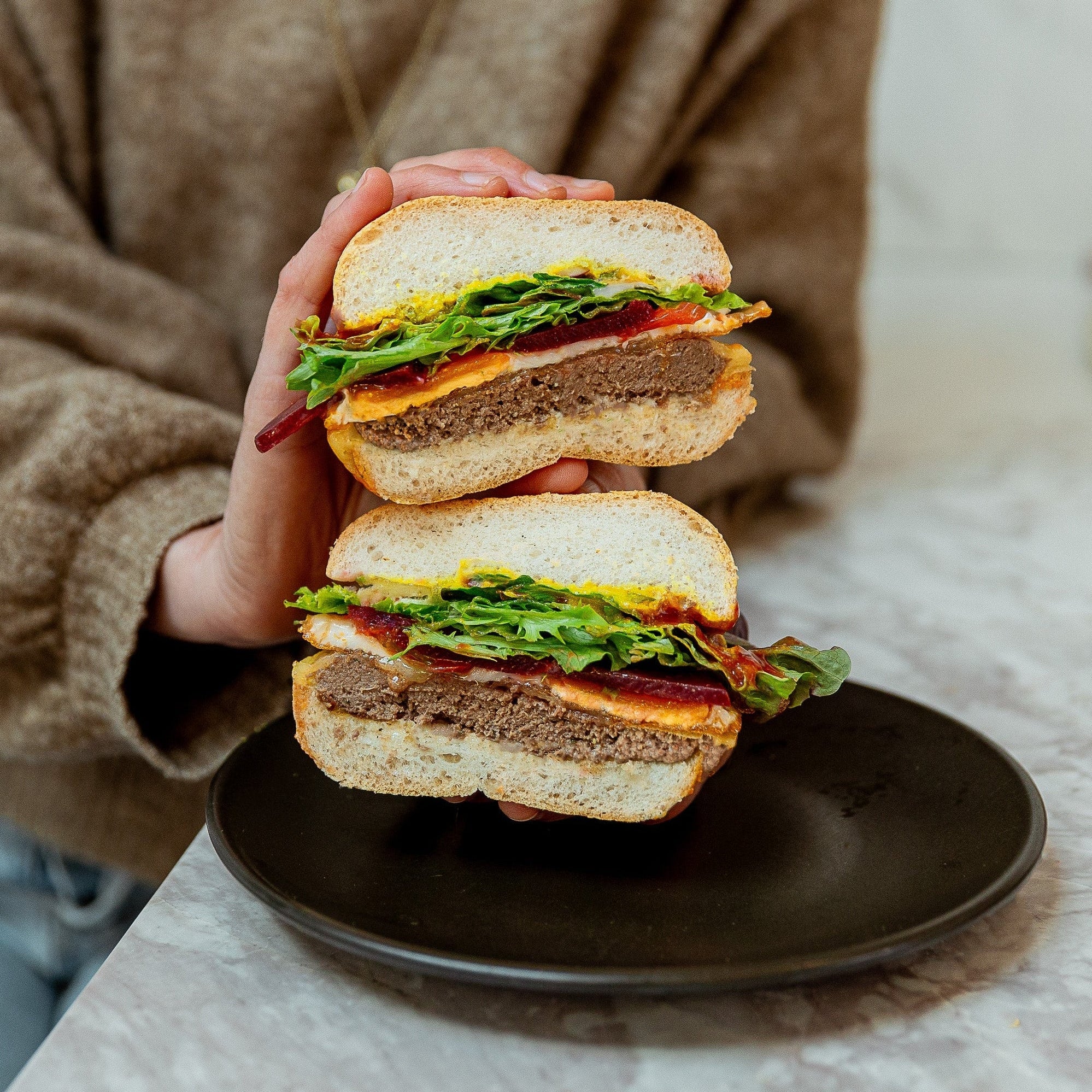 A person in a brown knit sweater holds a large cut burger in each hand over a black plate on a marble countertop. The sandwich has a thick brown patty, green lettuce, and red tomato slices.