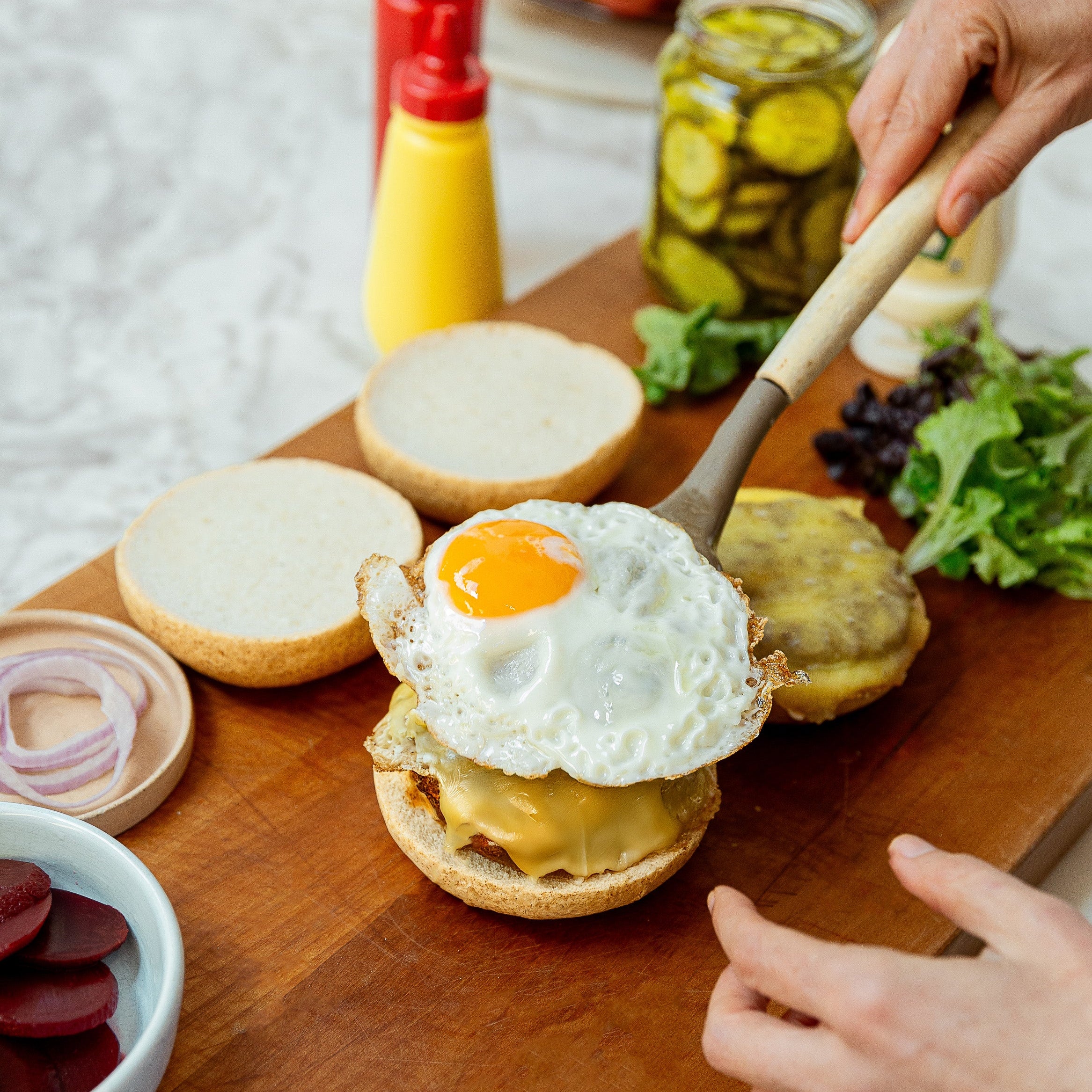 A close-up of a prepared Kiwi burger on a wooden cutting board, featuring a sunny-side-up egg on top of melted cheese and a beef patty, with hands adding the top bun. Condiments, sliced beets, and fresh greens are visible in the background.