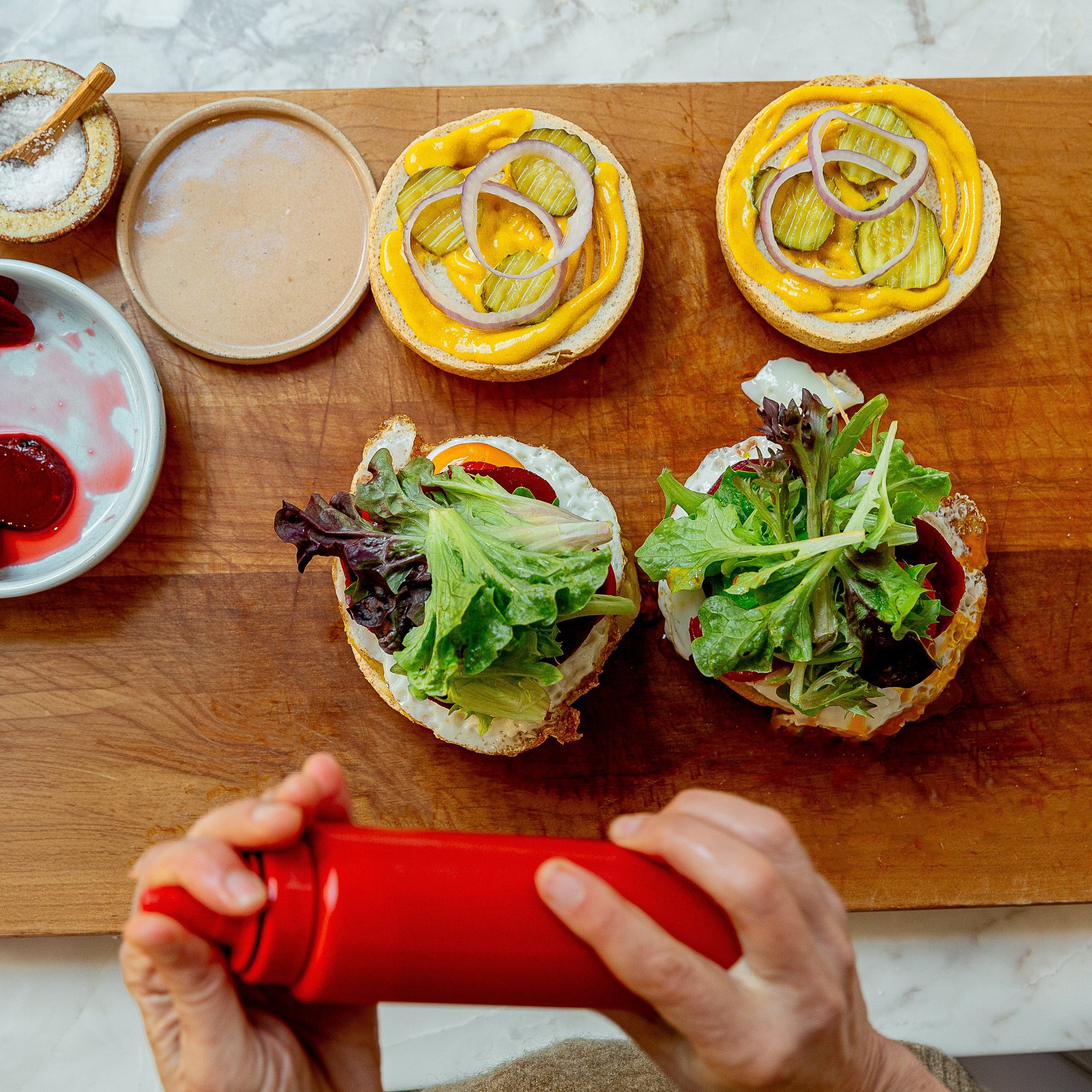 Overhead view of a wooden cutting board with two open burger buns topped with greens and pickles, surrounded by condiments and pickles. A person's hands are visible at the bottom holding a red squeeze bottle.