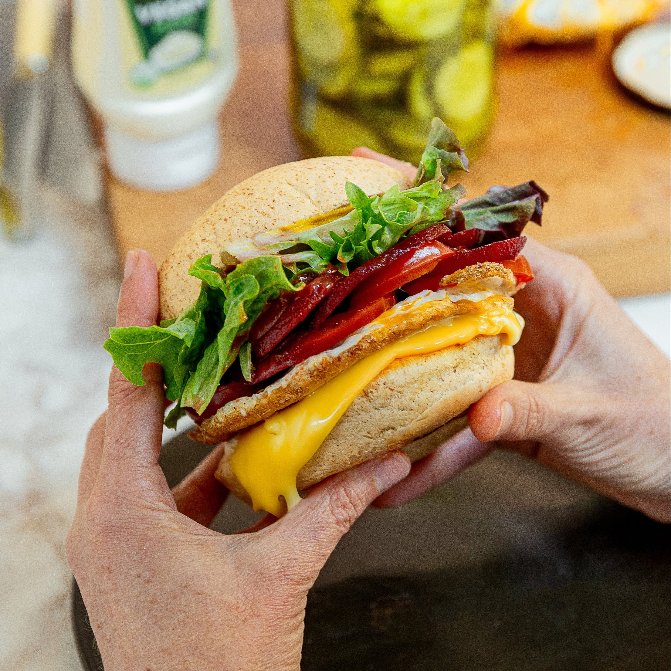 Two hands holding a kiwi burger filled with lettuce, beetroot, egg, beef patty, cheese and tomato with a jar of pickles and mayo in the background.