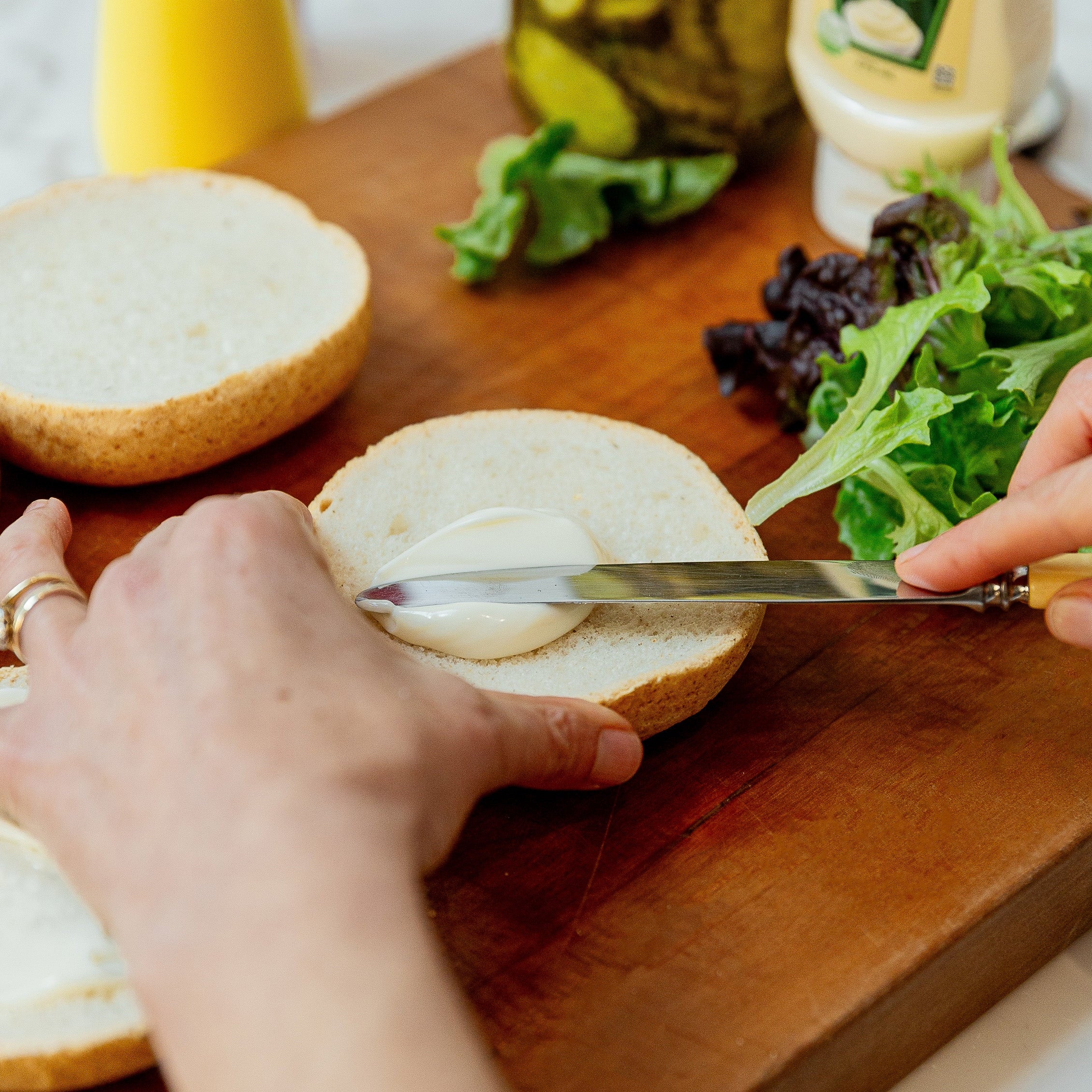 A close-up lifestyle shot of a person spreading vegan mayonnaise onto a burger bun on a wooden cutting board, with Heinz vegan mayonnaise and other ingredients visible in the background.
