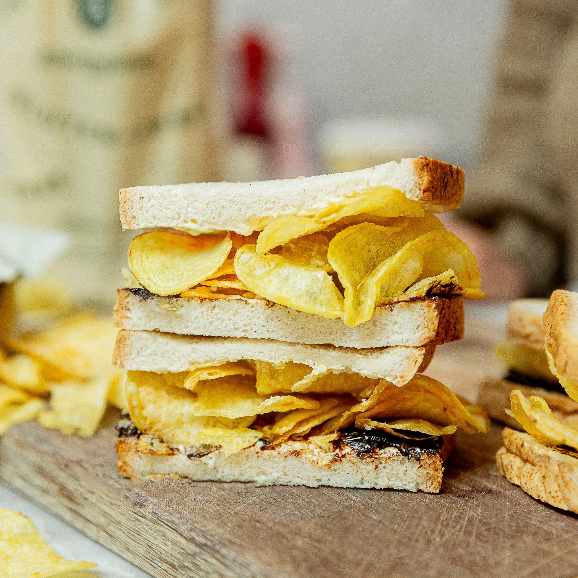 A stacked sandwich made with white bread and filled with crispy potato chips. and marmite resting on a wooden cutting board with scattered chips around it.