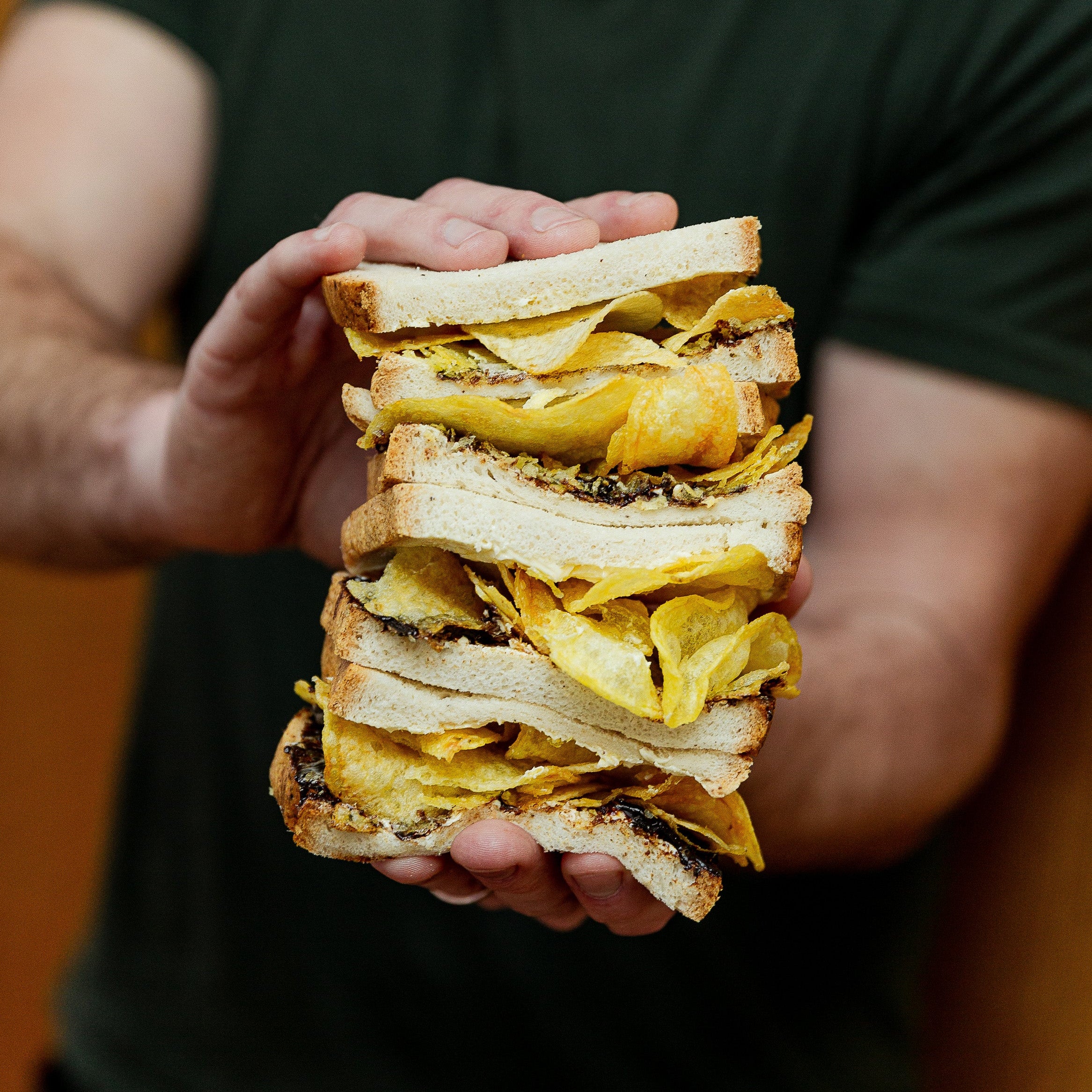 A person in a dark green t-shirt holds up a stacked sandwich made of white bread, crispy golden-yellow potato chips, and dark chocolate spread.