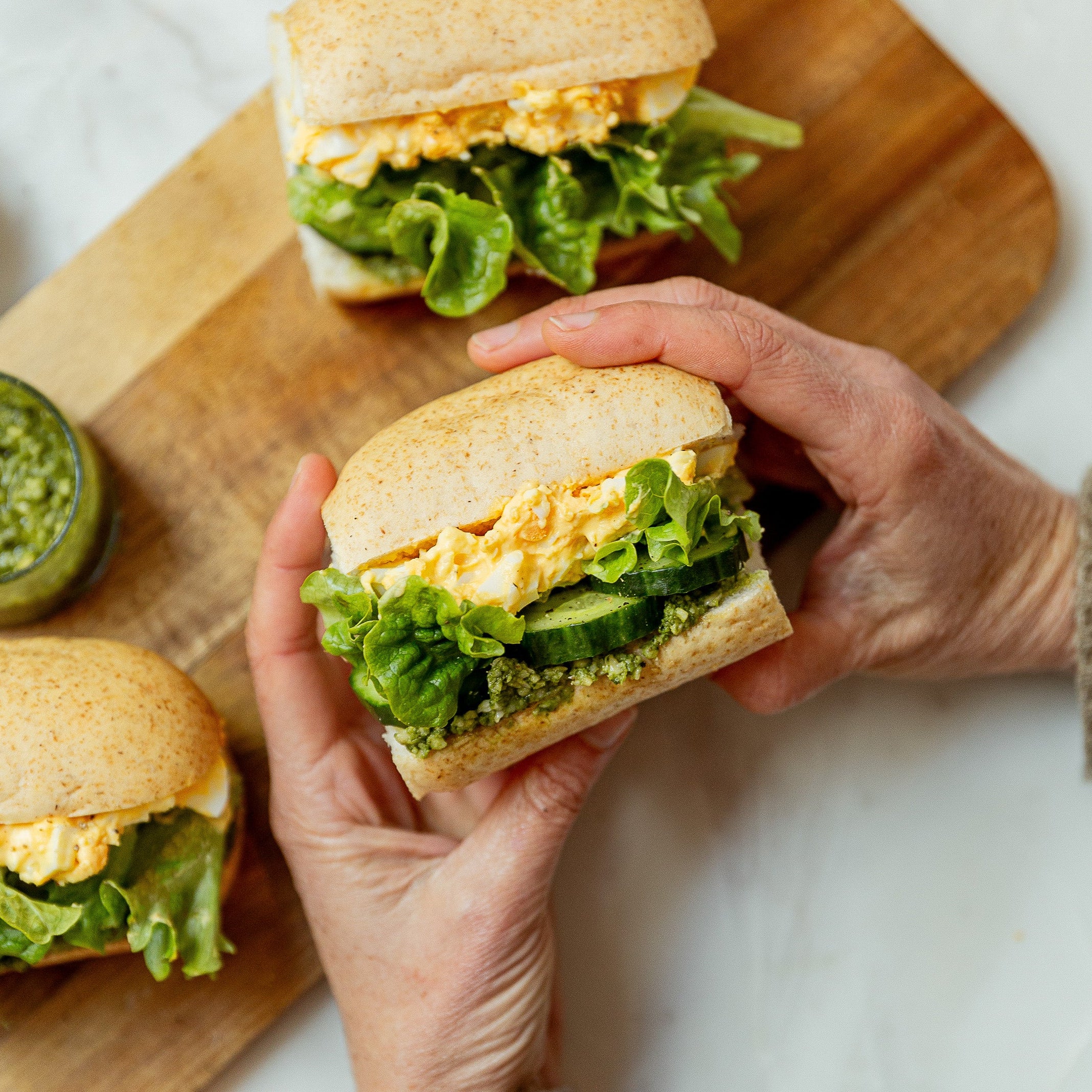 Two hands holding a small white roll filled with creamy egg, lettuce, green pesto and cucumber. A wooden board lays in the background with a jar of pesto and two more rolls.