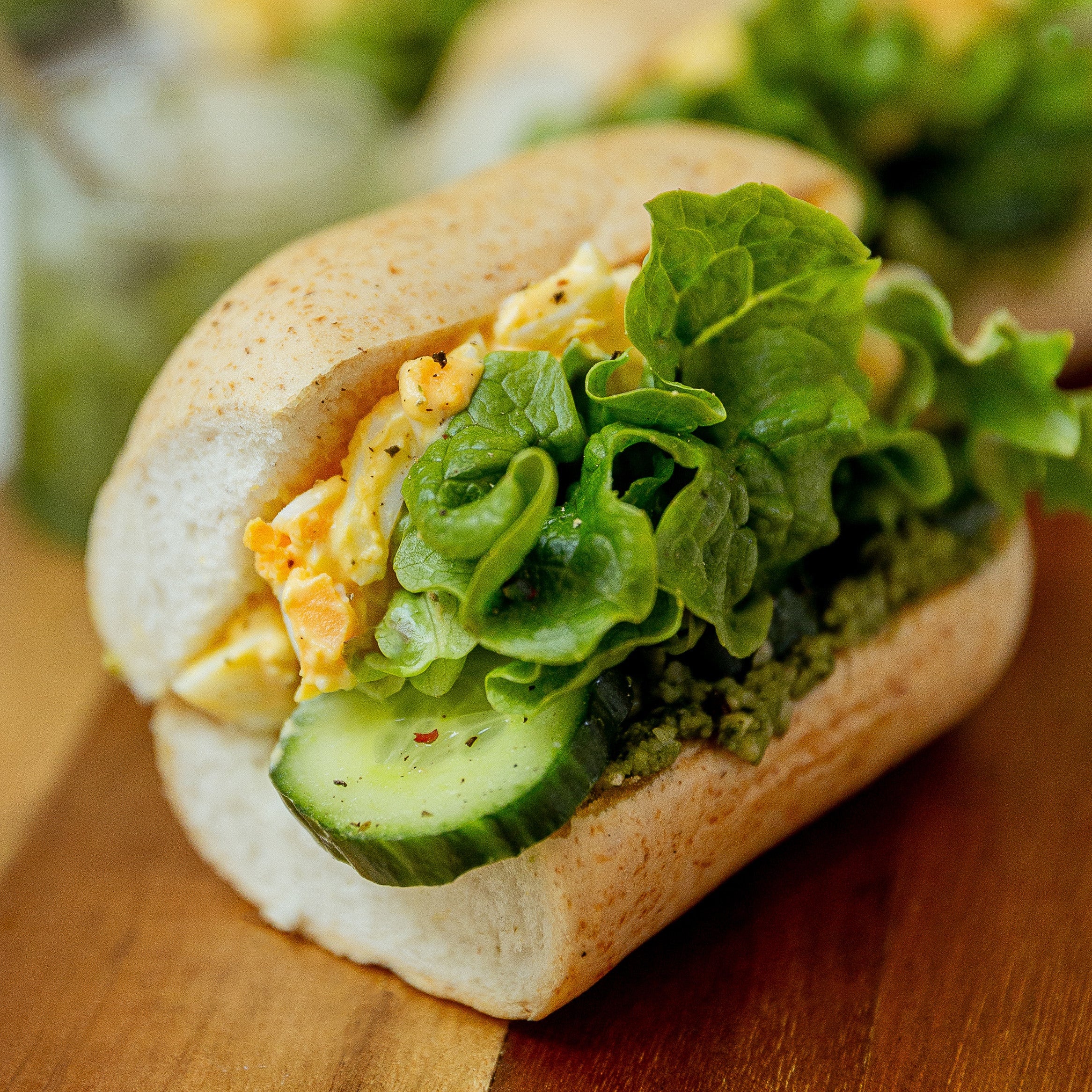 A close-up of a fresh sandwich on a wooden board, featuring a white roll filled with egg salad, crisp green lettuce, a cucumber slice, and a green spread, with additional blurred sandwiches in the background.