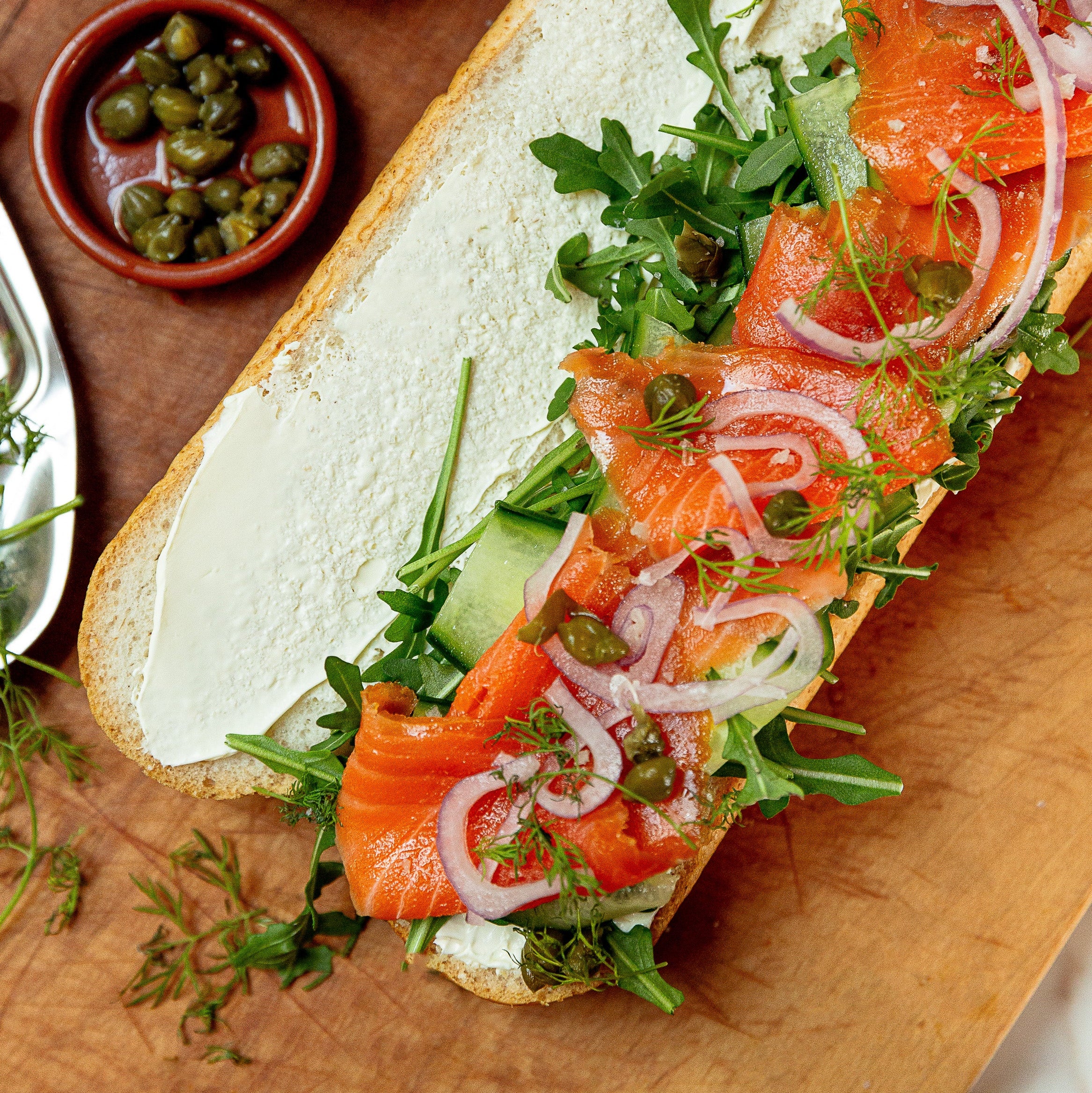 A long sandwich on a wooden board, filled with white spread, sliced salmon, arugula, red onion, and capers, with small bowls of garnish and a salt dish nearby.