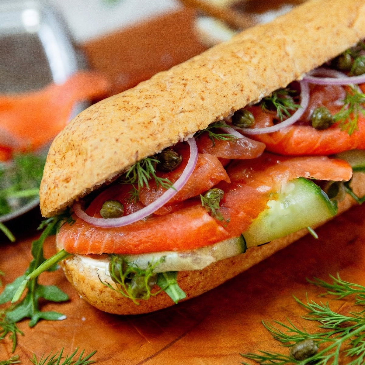 A close-up of a gourmet salmon sub sandwich on a wooden cutting board, filled with fresh salmon, cucumber, red onion, dill, and capers.