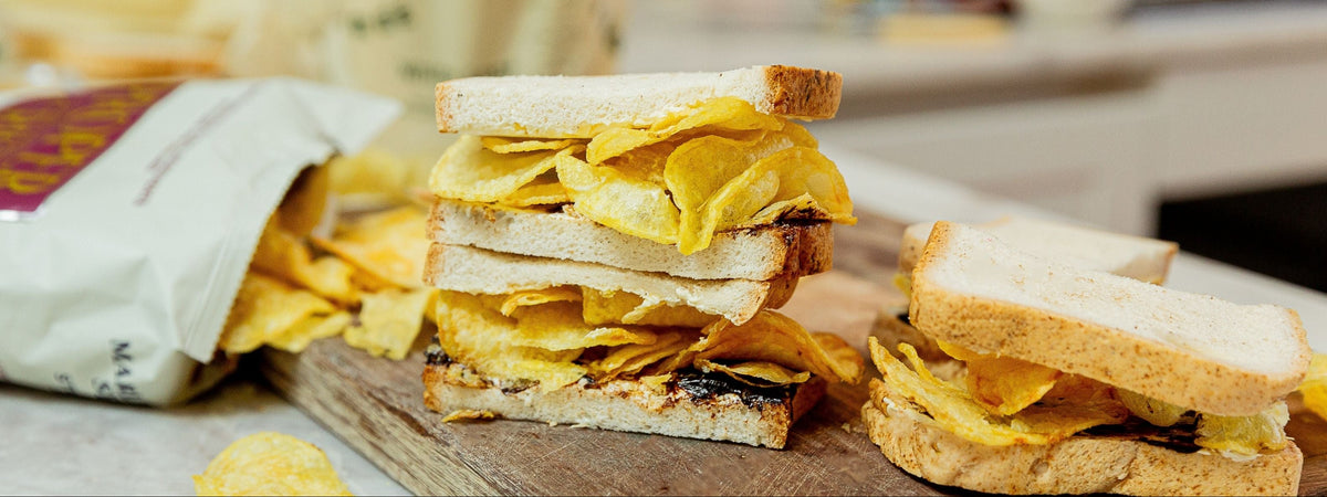 A close-up of a sandwich made with white bread and filled with potato chips, shown on a wooden board with a bag of chips in the background.